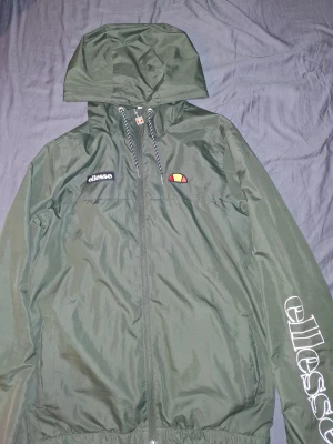 Grön vindjacka från Ellesse med huva - Snygg mörkgrön vindjacka från Ellesse med huva och dragkedja. Jackan har logga på bröstet och stort Ellesse-tryck på ena ärmen. Justerbar snörning i huvan och meshfoder på insidan. Perfekt för blåsiga dagar och streetwear-stil.