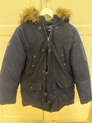 Mörkblå jacka från Polo Ralph Lauren - Snygg mörkblå parkas från Polo Ralph Lauren med fuskpäls på huvan och orange foder. Jackan har flera fickor med knappar, dragkedja och knäppning framtill samt patch på ärmen. Perfekt för kalla dagar och riktigt stilren look. Storleken är 14-16 i barn storlek som motsvarar ungefär XS. Skriv gärna vid frågor! Använd 2 gånger.