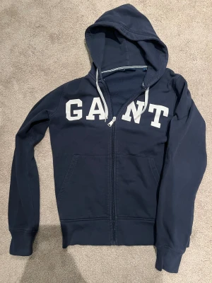 Marinblå hoodie från GANT med dragkedja - Snygg marinblå hoodie från GANT i storlek S med vit logotyp framtill. Tröjan har huva med snörning, praktiska fickor och hel dragkedja. Tillverkad i mjuk bomull som är skön att ha på sig. Perfekt för chill dagar och när du vill ha en clean look.