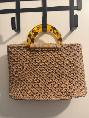 Beige handväska med leopardhandtag - Snygg beige handväska, handgjord virkad look och ett unikt handtag i transparent gul plast med leopardmönster. Väskan har en rektangulär form och är perfekt för dig som gillar trendiga detaljer och naturfärger.