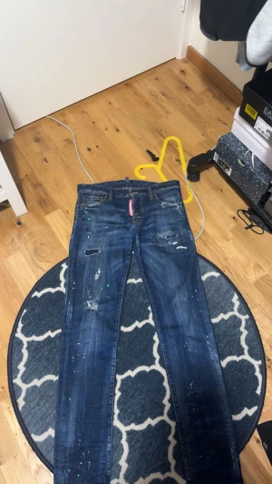 Blå jeans med slitningar och färgstänk - Säljer ett par blå jeans från Dsquared2 med coola slitningar och färgstänk i grönt och vitt. Jeansen har en röd logotyp-tag vid gylfen, klassisk femficksdesign och smal passform. Perfekt för dig som gillar en edgy streetstil.