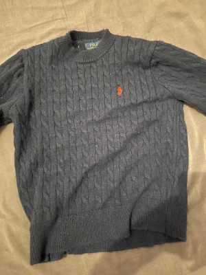 Mörkblå kabelstickad tröja Polo Ralph Lauren - Kabelstickad mörkblå tröja från Polo Ralph Lauren med klassisk rund hals och röd broderad logga på bröstet. Tröjan har ribbade muddar vid ärmslut och nederkant, perfekt för dig som gillar stilren och tidlös design.