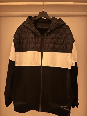 Svart och vit jacka/kofta Moose Knuckles - Snygg svart och vit jacka från Moose Knuckles med quiltad huva och axelparti. Jackan har dragkedja framtill, lång ärm och en cool blockdesign. Perfekt för dig som gillar streetwear och vill sticka ut med stilren look.