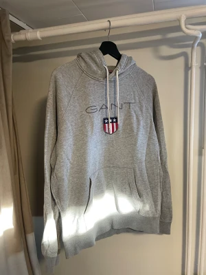 Gant Hoodie – Grå, Storlek L - Säljer en grå Gant hoodie i storlek L. Hoodien är i gott begagnat skick med spår av användning, men fortfarande mjuk, bekväm och fullt användbar. Klassisk Gant-design med broderad logotyp och sköld på bröstet.