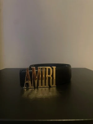 Svart skärp med gulddetalj - Snyggt svart bälte med en stor, glänsande guldfärgad logotyp som spänne. Bältet är gjort i skinn med en stilren och klassisk design som sticker ut med den ikoniska bokstavsspännet framtill.