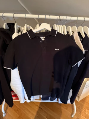Svart pikétröja från BOSS med vit kant - Svart pikétröja från BOSS i storlek 16-M sitter som S. Med vit kant på krage och ärmslut. Klassisk design med tre knappar framtill och BOSS-logga broderad på bröstet. Perfekt för dig som gillar stilrena och snygga plagg med premiumkänsla.