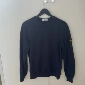 Mörkblå sweatshirt från Stone Island - Snygg mörkblå sweatshirt från Stone Island Junior med klassisk rund hals och lång ärm. Tröjan har den ikoniska Stone Island patchen på vänster ärm och är gjord i mjuk bomull. Perfekt för dig som gillar streetwear och vill ha en clean look.