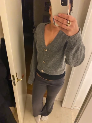 Grå kofta från H&M med guldfärgade knappar - Mysig grå kofta från H&M i storlek M. Koftan är stickad och har en croppad passform med v-ringad hals. Enkel och stilren design som funkar till många outfits.