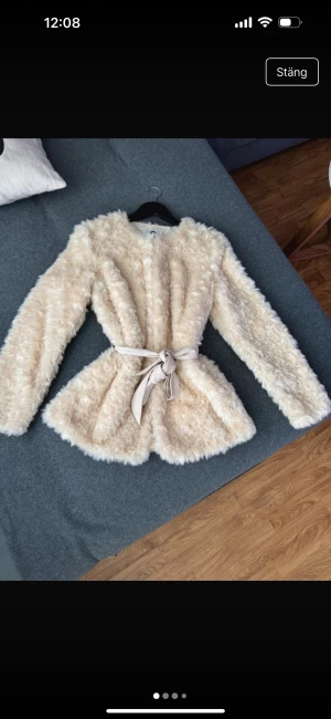 Beige pälsjacka med bälte - Supermjuk och fluffig beige pälsjacka med markerad midja tack vare ett brett knytbälte. Jackan har lång ärm, rund halsringning och är helfodrad med slät insida. Perfekt för dig som vill ha en cozy men ändå stylish look.