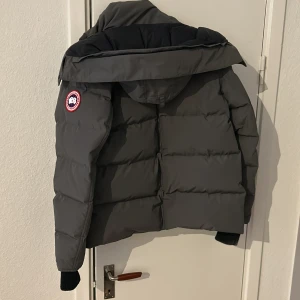 Mörkgrå dunjacka från Canada Goose - Säljer en mörkgrå dunjacka från Canada Goose med stor huva och avtagbar pälsdetalj. Jackan har klassisk pufferstil, ribbade muddar och ikonisk logga på ärmen. Perfekt för kalla vinterdagar och riktigt snygg streetstil.