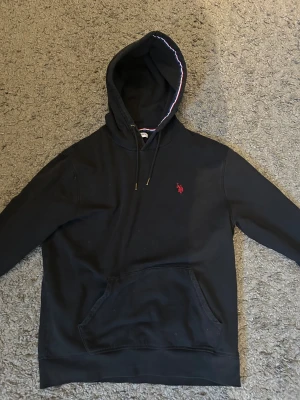 Svart Polo hoodie  - Svart Polo hoodie med röd broderad logga på bröstet. Tröjan har huva med snörning och en klassisk känguruficka framtill. Insidan av huvan har en snygg randig detalj i vitt och rött. Perfekt för chill dagar. Skriv för mer bilder 
