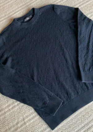 Stickad tröja Dressmann - Snygg mörkblå stickad tröja från Dressmann i storlek M. Tröjan har ett diskret strukturmönster och rund hals med ribbade muddar vid ärmslut och nederkant. Perfekt för dig som gillar stilrena och klassiska plagg med lite extra detalj.