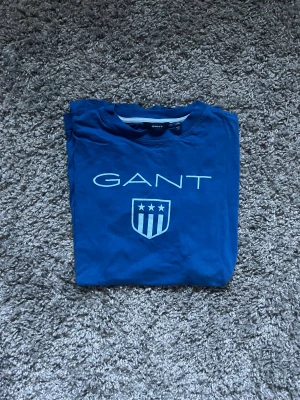 Blå GANT t-shirt med tryck - Snygg blå t-shirt från GANT med stort vitt logotryck och sköld på bröstet. Klassisk rund hals och korta ärmar. Tillverkad i mjuk bomull som är skön mot huden. Perfekt för dig som gillar stilrena märkesplagg med sportig vibe.