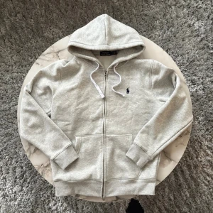 Polo Ralph Lauren kofta / zip up - Snygg ljusgrå hoodie från Polo Ralph Lauren med dragkedja och vit snörning i huvan. Tröjan har det klassiska blå Polo-logot på bröstet, två fickor fram och ribbade muddar. Perfekt för chill dagar och enkel att matcha med jeans eller joggers. Väldigt bra och högkvalitativ material som gör koftan väldigt skön att ha på sig. 