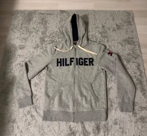 Grå hoodie Tommy Hilfiger - Snygg grå hoodie från Tommy Hilfiger med mörkblå broderad logga på bröstet och klassiska Hilfiger-flaggan på ärmen. Tröjan har dragkedja, huva med vita snören och två fickor framtill. Perfekt för chill dagar och streetstyle.