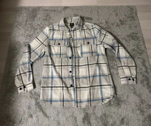 Rutig skjorta från H&M i grått och blått - Säljer en snygg rutig skjorta från H&M i storlek M. Skjortan har en cool färgkombination med grå, vit och blå toner, klassisk krage och svarta knappar. Två bröstfickor med knappstängning ger extra stilpoäng. Perfekt för dig som gillar avslappnad och trendig look.