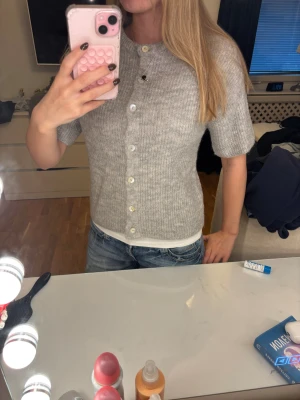 Stickad kofta - Säljer en grå stickad kofta från Gina Tricot i ullblandning. Koftan har korta ärmar, rund hals och knappar hela vägen framtill. Ribbad struktur som ger en mysig och chill vibe. Perfekt att slänga på över en t-shirt eller linne.