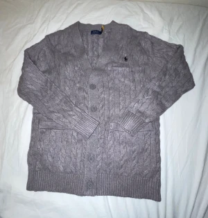 Grå kofta från Polo Ralph Lauren - Snygg grå kofta från Polo Ralph Lauren i storlek S. Den har kabelstickat mönster, ribbade muddar och knappar framtill. Använd men inget som syns, köpt för ungefär 2500kr. 90% ull, 10% Kashmir.
