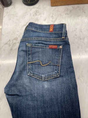 Blå jeans från 7 For All Mankind - Snygga blå jeans från 7 For All Mankind med klassisk femficksdesign och gul kontrastsöm på bakfickan. Jeansen har en normal passform och är tillverkade i mjukt denimtyg med röda detaljer på etiketten.