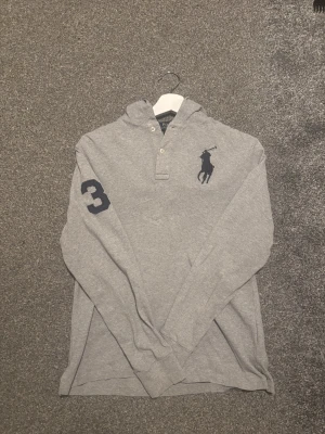 Grå henley från Polo Ralph Lauren - Säljer en grå långärmad henley från Polo Ralph Lauren med klassisk krage, knappar vid halsen och stor marinblå logga på bröstet. Snygg detalj med en mörkblå siffra 3 på ärmen. Perfekt för en chill och stilren look. Storlek 14-16