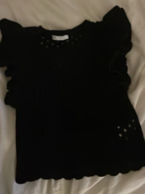 Svart stickad topp med volangärm - Säljer en svart stickad topp med korta volangärmar och rund halsringning. Toppen har ett diskret hålmönster och vågig kant nedtill som ger en söt touch. Perfekt att styla med jeans eller kjol för en trendig look.