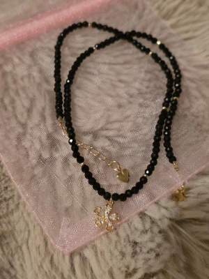 Handgjort halsband - Elegant handgjort halsband med små svarta fasetterade pärlor och guldfärgade detaljer. I mitten sitter en fin blomberlock med gnistrande stenar som ger ett stilrent men feminint uttryck.Längd: 38 cm Passform: Sitter vid nyckelbenet (kort halsband) Skick: Nytt, handgjort
