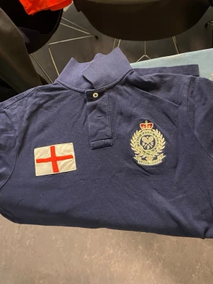 Marinblå Ralph Lauren England piké - Säljer en marinblå pikétröja från Polo Ralph Lauren med broderad England-flagga och tennismärke på bröstet. Baksidan har stora bokstäver med 'ENGLAND' sopatchat. Klassisk krage med två knappar och korta ärmar. Materialet är mjuk bomull och modellen är custom fit.