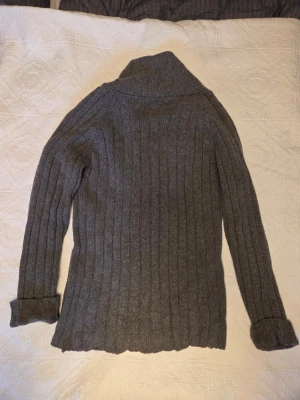 Grå ribbad kofta från Filippa K - Säljer en grå ribbstickad kofta från Filippa K med bred krage och fyra knappar framtill. Koftan är i en mix av bomull och lammull, har långa ärmar och en klassisk, lite längre passform. Perfekt för lager på lager under kyliga dagar.