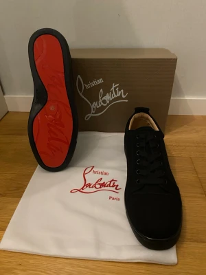 Christian Louboutin Skor Helt Nya!!! - Säljer ett par svarta sneakers från Christian Louboutin med ikonisk röd sula. Skorna har rund tå, snörning och är tillverkade i mocka med detaljer i skinn. Kommer med originalkartong, dustbag och äkthetskort. Perfekta för dig som vill ha något extra till din outfit.                                                                        Helt Nya!!!