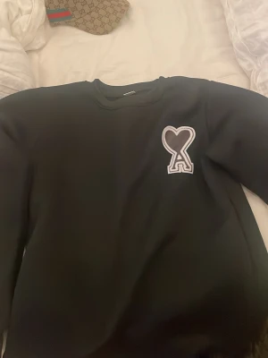 Svart sweatshirt från Ami Paris - Säljer en svart sweatshirt från Ami Paris med det ikoniska hjärta-A-märket broderat på bröstet. Tröjan har rund halsringning och lång ärm, perfekt för en clean och stilren look.