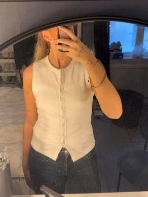 Vit ribbad linnetopp från Edikted - Säljer en vit ribbad linnetopp från Edikted i storlek M. Toppen är ärmlös med rund halsringning och knappar hela vägen framtill. Perfekt att styla med jeans eller kjol för en clean look.