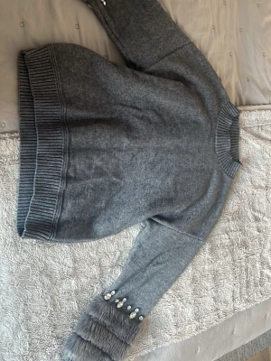 Grå stickad tröja med pälsdetalj - Säljer en grå stickad tröja från Cliche' i storlek M. Tröjan har ribbade muddar och krage, samt långärmade ärmar med fluffig pälsdetalj och dekorativa pärlor vid ärmslutet. Perfekt för dig som gillar unika detaljer och vill ha något extra i garderoben.