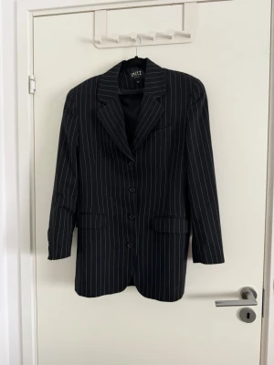 Svart randig kavaj från IMITZ - Snygg svart kavaj från IMITZ med tunna vita ränder. Klassisk modell med tre knappar framtill, krage och två fickor med lock. Långärmad och med en rak passform som ger en stilren look. Perfekt för dig som vill ha en tidlös och trendig kavaj.