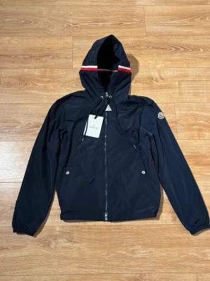 Marinblå vindjacka från Moncler - Snygg marinblå vindjacka från Moncler med huva och vita/röda detaljer. Jackan har dragkedja framtill, snörning i huvan och två fickor med tryckknappar. Moncler-logga på ärmen och lätt, sportig känsla. Perfekt för dig som gillar stilren och funktionell design.