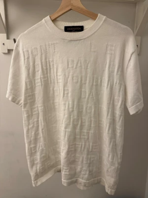 Vit Louis Vuitton t-shirt med textur - Exklusiv vit t-shirt från Louis Vuitton med upphöjd text över hela plagget. Klassisk rund halsringning och korta ärmar. Snygg detalj med Louis Vuitton Paris-märke i nacken. Tillverkad i mjuk bomull som känns lyxig och skön mot huden.