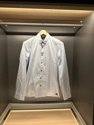 Ljusblå skjorta från Selected Homme - Snygg ljusblå skjorta från Selected Homme med klassisk krage och svarta knappar. Insidan av kragen har ett rutigt tyg i brunt och blått som ger en cool detalj. Skjortan är långärmad och gjord i bomull för en skön känsla.