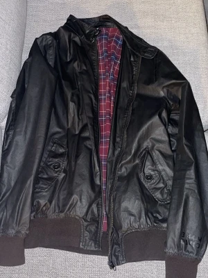 Svart Vintage bomberjacka med rutigt foder - Säljer en Vintage svart bomberjacka med rutigt foder i vinrött och blått. Jackan har dragkedja framtill, två fickor med knapp och ribbade muddar vid ärmar och midja. Materialet är syntet och jackan har en klassisk, loose passform.