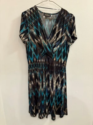 Mönstrad kortklänning från Alice Bizous - Snygg kortklänning med v-ringning och omlott-effekt från Alice Bizous. Klänningen har ett grafiskt zigzag-mönster i svart, blått och beige, samt korta ärmar. Tillverkad i ett mjukt och följsamt polyestertyg som ger en skön passform.
