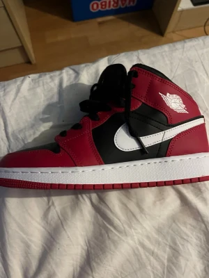 Nike Air Jordan 1 Mid Svart/Röd - Säljer ett par Nike Air Jordan 1 Mid i svart och rött med vita detaljer. Skorna har klassisk snörning, perforerad tåbox och den ikoniska swoosh-loggan på sidan. Tillverkade i syntet och skinn för en clean och sportig look.