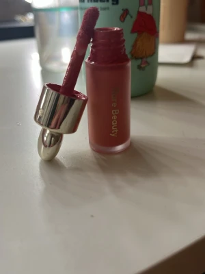 Rare Beauty flytande rouge Happy - Säljer en flytande rouge från Rare Beauty i nyansen Happy. Färgen är en fräsch rosa med kall underton och kommer i en matt glasflaska med silvrigt lock och applikator. Perfekt för att få ett naturligt rosigt glow på kinderna.