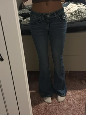 Blå bootcut jeans med broderade fickor - Säljer ett par blå bootcut jeans med snygga broderade detaljer på bakfickorna. Jeansen har låg midja och klassisk femficksdesign. Materialet är jeans och passformen är loose med utsvängda ben. Perfekta för dig som gillar retrovibbar och vill sticka ut lite extra.