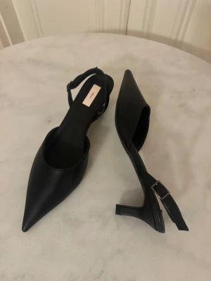 Svarta pumps med spetsig tå och låg klack - Säljer ett par stilrena svarta pumps från Nelly  i storlek 41. Skorna har en spetsig tå, låg klack och en smal rem med spänne runt. Eftersom man kan spänna de så skulle det passa någon som har storlek 40 också. 