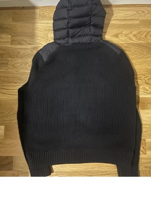 Svart Moncler Cardigan - Säljer en svart pufferjacka från Moncler med stickade ärmar och detaljer. Jackan har huva, dragkedja framtill och Moncler-logga på ärmen. Perfekt för dig som vill ha en varm och snygg jacka med både quiltad och stickad känsla. Hör av dig för mer frågor osv!