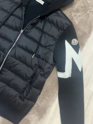 Svart Moncler pufferjacka med huva - Säljer en svart pufferjacka från Moncler med huva och dragkedja framtill. Jackan har stickade ärmar med vit zigzagdetalj och Moncler-logga på armen. Kroppen är quiltad och fylld med dun, medan ärmarna är i mjukt material. Perfekt för dig som vill ha en snygg och sportig look.