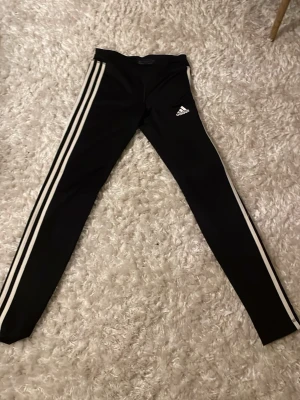 Svarta Adidas leggings med vita ränder - Säljer ett par svarta Adidas leggings med klassiska vita ränder längs sidorna och Adidas-logga på låret. De är tighta och har en sportig look, perfekta för träning eller chill. Gjorda i stretchigt syntetmaterial för maximal komfort. Storlek 164