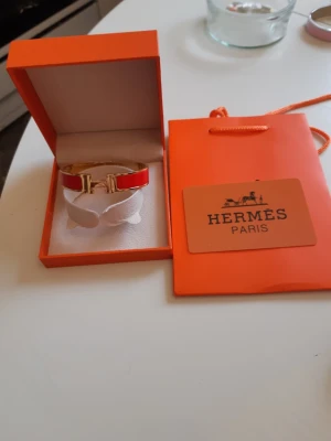 Hermes armband  - Bilderna visar ett Hermès-armband i röd och guld färg med tillhörande ask och påse, 