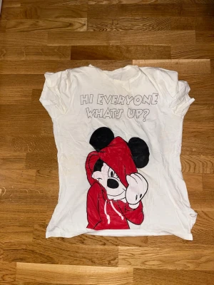 Vit t-shirt med Mickey Mouse tryck - Vit t-shirt med ett stort tryck av Mickey Mouse i röd hoodie och texten 'HI EVERYONE WHAT'S UP?' på bröstet. T-shirten har korta ärmar och en klassisk rund hals. Perfekt för dig som gillar Disney och vill ha en chill och lekfull stil.