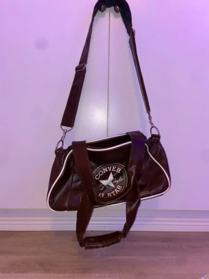 Brun axelväska Converse All Star - Säljer en brun axelväska från Converse All Star med vit piping och stort logotryck på sidan. Väskan har justerbar axelrem, dubbla handtag och dragkedja upptill. Insidan är svart med flera fack för småprylar. Mått: längd, ca 44 cm. Höjd: ca 20 cm