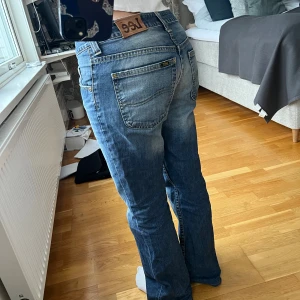 Blå bootcut jeans från Lee - Säljer ett par klassiska blå jeans från Lee med bootcut passform och låg midja. Jeansen har ljusare partier framtill för en snygg vintage-look. Tillverkade i jeansmaterial med fem fickor och Lee-lapp bak i midjan. Osäker på exakt storlek men passar mig som är 165 cm och ofta bär storlek S i jeans. Skriv privat för mått. 