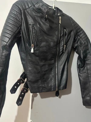 Svart skinnjacka med dragkedjor - Säljer en svart skinnjacka med coola detaljer som silvriga dragkedjor, quiltade axlar och två spännen i midjan. Jackan har en asymmetrisk dragkedja framtill och flera fickor med dragkedja. Perfekt för dig som vill ha en edgy look.